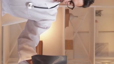Vertical video. Clinical diagnosis. Male traumatic surgeon. Study information. Smart man looking x-ray photo making notes on paper sitting light office room interior.