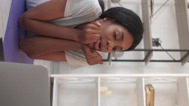 Vertical video. Lazy sport. Relaxed woman. Online chat. Happy casual lady in activewear talking laptop after workout on yoga mat light room interior.