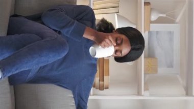 Vertical video. Morning coffee. Happy woman. Enjoying moment. Smiling dreamy lady drinking delicious beverage resting on couch light room interior.