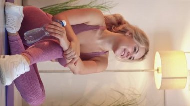 Vertical video. Sporty woman. Healthy lifestyle. Home fitness. Positive sportswoman in activewear sitting on yoga mat holding water bottle light room interior.