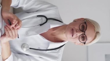 Vertical video. Medical consultation. Female doctor. Online visit. Middle-aged woman in spectacles greeting talking on camera in light room interior.