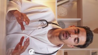 Vertical video. Online conference. Male doctor. Medical support. Inspired man in whites talking on camera sitting work desk in light room interior.