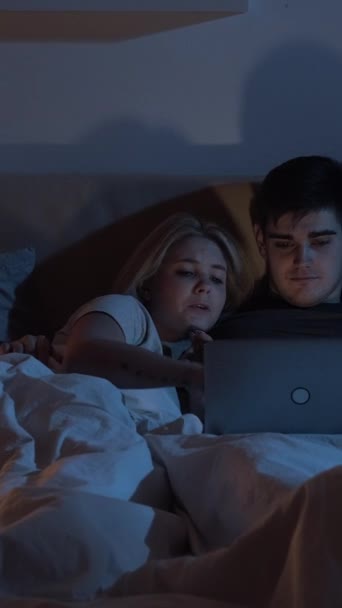 夜のオンラインショッピング カップルの購入 インターネット消費主義 興奮した女性は暗い寝室の遅いベッドでノートパソコンを使用してギフトを購入する思いやりの夫を物乞い 垂直ビデオ — ストック動画