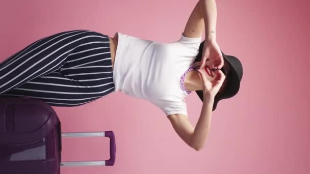 垂直ビデオ 愛を送る インスピレーションを受けた女性旅行者 休暇の喜び かなり幸せな女性夏ルックスポージングとともにスーツケースショーハートサイン上のピンクの背景 — ストック動画