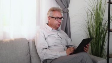 Video conference. Virtual meeting. Working from home. Relaxed confident senior business man in glasses discussing project using tablet on couch in light living room interior.