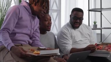 Online sport. Happy male friends. Fast food dinner. Positive black men talking looking video on laptop eating pizza in light room interior.