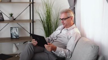 Family video call. Virtual chat. Online communication. Relaxed cheerful middle-aged man in glasses waving hi on tablet camera sitting on couch in light home living room interior.