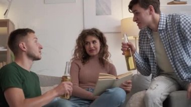 Different interests. Happy friends. Home leisure. Inspired two men cheering with beer while blaming woman reading book on sofa light room interior.