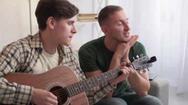 Music joy. Male player. Home leisure. Inspired casual man playing wonderful song on guitar for amazed friend sitting sofa in light room interior.