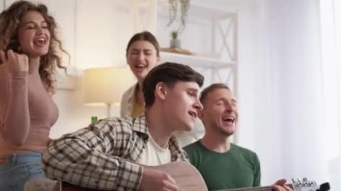 Home party. Meeting friends. Life music joy. Happy men and women enjoying time together playing guitar singing and dancing in light room interior.