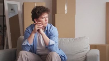 Move house. Upset man. New life sadness. Disappointed guy feeling sadness and grief sitting sofa with packed cardboard boxes light room interior.