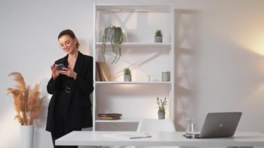 Work break. Happy woman. Mobile communication. Smiling elegant lady in suit chatting in smartphone staying light office room interior copy space.