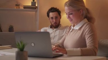 Interesting project. Happy colleagues. Late work. Inspired woman showing information on laptop to relaxed man sitting sofa drinking coffee in light room interior.