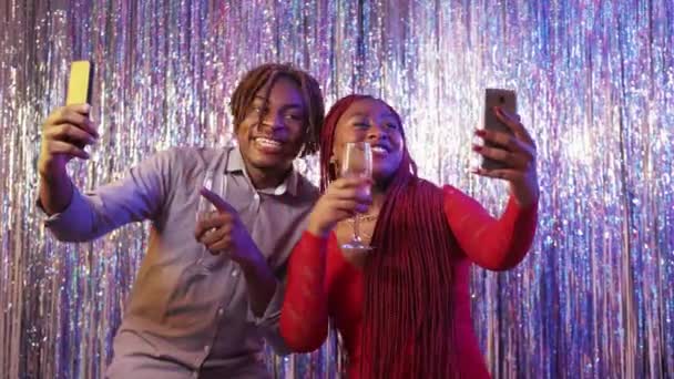 网上问候 兴奋的夫妇 节日庆祝活动 快乐的黑人男人和女人在手机上拿着香槟 摆出闪光的瀑布幕背景 — 图库视频影像