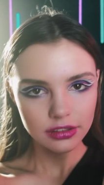 Vertical video. 90s makeup. Retro fashion. 2000s night look. Smiling confident teen girl face with blue color eyeshadow pink lips artistic visage posing in neon light on dark.