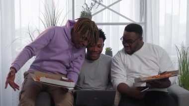 Online meeting. Black men. Home dinner. Happy male friends eating pizza during virtual conference on laptop light room interior.