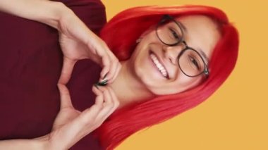 Vertical video. Heart gesture. Love affection. Romantic message. Portrait of cute cheerful smiling girl with red hair showing sympathy hand sign on orange background.