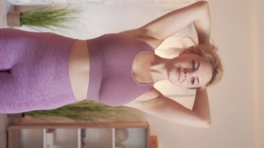 Vertical video. Fitness preparing. Pretty woman. Home sport. Cheerful sporty lady in sportswear tying hair in ponytail before exercise light room interior.