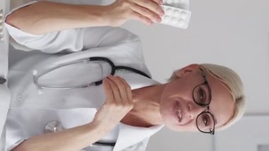 Vertical video. Bad medicine. Female doctor. Online consultation. Middle-aged woman in whites throwing away tablets sitting light room interior.