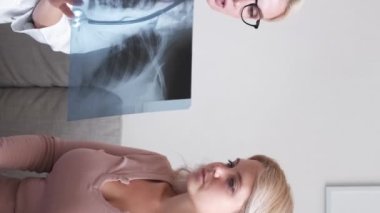 Vertical video. Lung fever. Female doctor. Labs result. Medical woman showing x-ray photo consulting patient sitting sofa light office room interior.