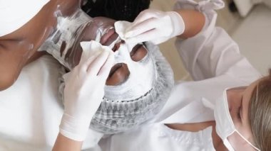 Vertical video. Clinical cosmetology. Female beauty. Salon spa. Professional cosmetologist removing facial mask from woman face skin light room interior.