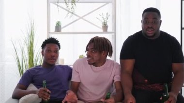 Friendly meeting. Black men. Interesting movie. Relaxed hipster guys talking together with beer bottles sitting sofa light room interior.