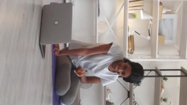 Vertical video. Remote fitness. Happy woman. Online connection. Friendly slim lady in sportswear talking on laptop having virtual call light room interior.