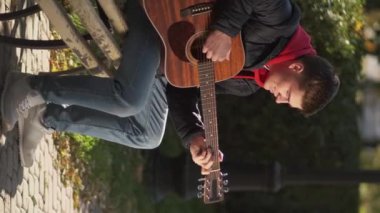 Vertical video. Street player. Male singer. Enjoying autumn. Inspired happy man playing melody on guitar sitting bench in park.