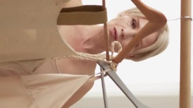 Vertical video. Elegant look. Middle-aged woman. Festive preparation. Beautiful puzzled lady choosing dress on holiday party in light room interior.