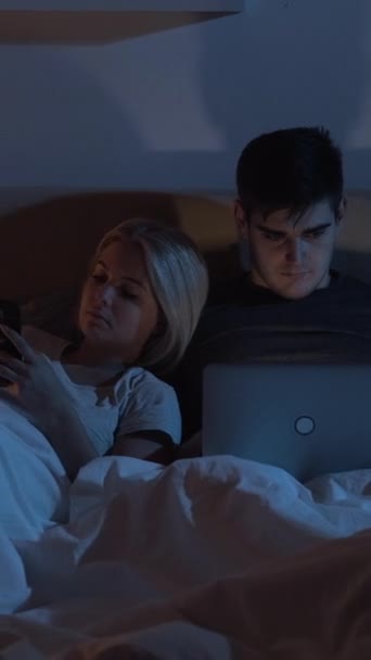 小夜叉 数码夫妻 晚了上网 在黑暗的卧室里 女人用手机丈夫在笔记本电脑上上网工作 垂直录像 — 图库视频影像