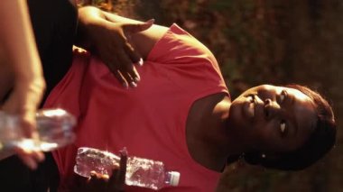 Vertical video. Fitness rest. Workout break. Outdoor communication. Cheerful relaxed female friends with water bottles talking discussing healthy lifestyle in nature park.