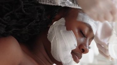 Vertical video. Facial cosmetic. Beauty salon. Skincare procedure. Unrecognizable dermatologist applying white clay mask woman face skin in clinic.