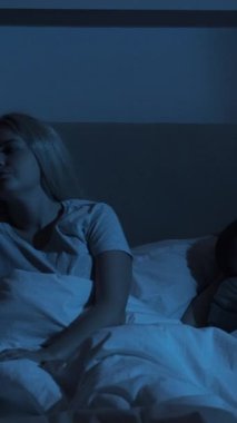 Gece uykusuzluğu. Uyku bozukluğu. Yorgunluk endişesi. Yorgun ve sıkılmış bir kadın karanlık bir odada huzurlu bir kocayla yatakta oturuyor. Dikey video.