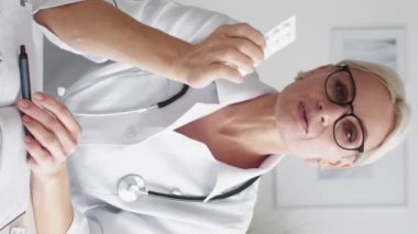 Vertical video. Medicine consultation. Female doctor. Online support. Middle-aged woman in whites consulting about tablets sitting light room interior.