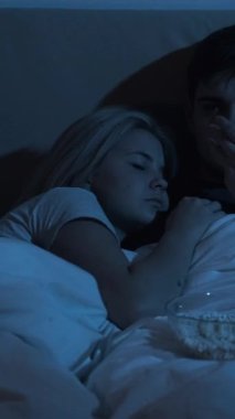 Gece filmi. Uykusuzluk sorunu. Geç saatlere kadar boş vakit. Sıkılmış koca TV izlerken yatakta patlamış mısır yerken yorgun kadın karanlık odada uyuyor. Dikey video.