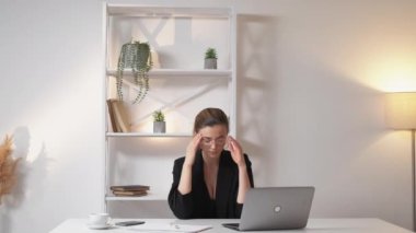 Headache suffer. Tired woman. Hard work. Exhausted elegant lady massaging head temples working laptop in light office room interior.