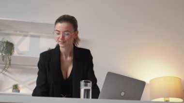 Office work. Elegant woman. Digital technology. Smiling lady in suit checking documents typing laptop in light room interior.