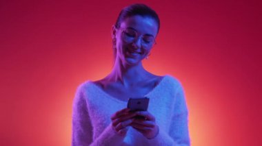 Mobile communication. Happy woman. Neon light portrait. Joyful pleased lady typing smartphone posing blue red background.