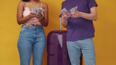 Vacation budget. Rich couple. Journey spending. Unrecognizable man and woman counting cash dollars posing with suitcase yellow background.