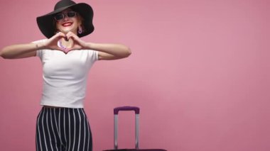 Sending love. Inspired woman. Travelling joy. Pretty happy lady in elegant summer look dancing with suitcase showing heart sign on pink background copy space.