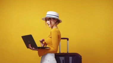 Online tickets. Excited woman. Vacation planning. Positive inspired lady in hat searching on laptop posing with suitcase on yellow background.