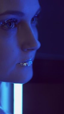 Vertical video. Glitter makeup. Neon girl. Nightclub fashion. Blue purple color flash light woman face with sparkling metallic eyeshadow lips in mirror on dark.