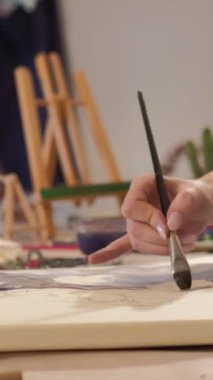 Vertical video. Painting hobby. Art inspiration. Creative talent. Closeup of female artist hand using brush creating watercolor artwork on canvas at workplace.