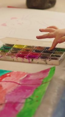 Vertical video. Finger painting. Kid art. Young artist. Child hand mixing colors in palette sprinkling watercolor paint over abstract artwork.