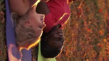 Vertical video. Fitness rest. Workout break. Park leisure. Happy diverse female friends talking chilling after training on yoga mat on fall grass.