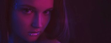 Neon face. Sparkling makeup. Disco vogue. Night portrait of purple blue color light woman with shimmering glitter skin metallic eyeshadow lips on dark black copy space background.