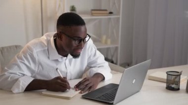 Corporate learning. Business lesson. Online education. Happy man student writing notes watching laptop sitting desk home workspace.