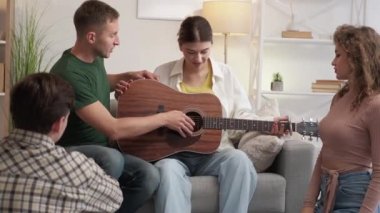 Guitar lesson. Supportive friends. Home meeting. Inspired boyfriend trying to teach playing girlfriend on acoustic instrument in light room interior.
