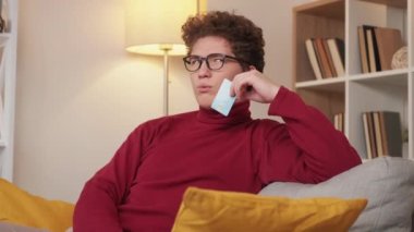 Purchase considering. Puzzled man. Credit money. Pensive guy sitting sofa with plastic card thinking about costs payment in light room interior.