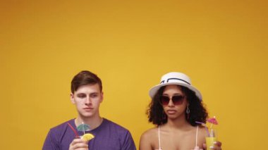 Summer tour. Conspirator couple. Odd situation. Weird man and woman in hat sunglasses looking each other posing with cocktails on yellow background.